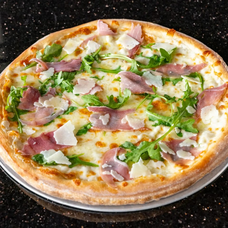 White pizza with prosciutto, arugula, and shaved Parmesan cheese on a dark speckled surface.