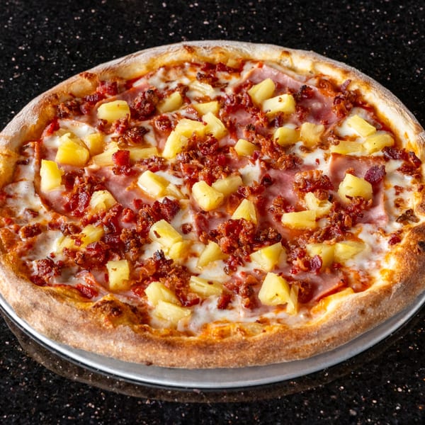 Grande Pizza's Hawaiian pizza, topped with ham, bacon, pineapple, and melted cheese on a dark surface.