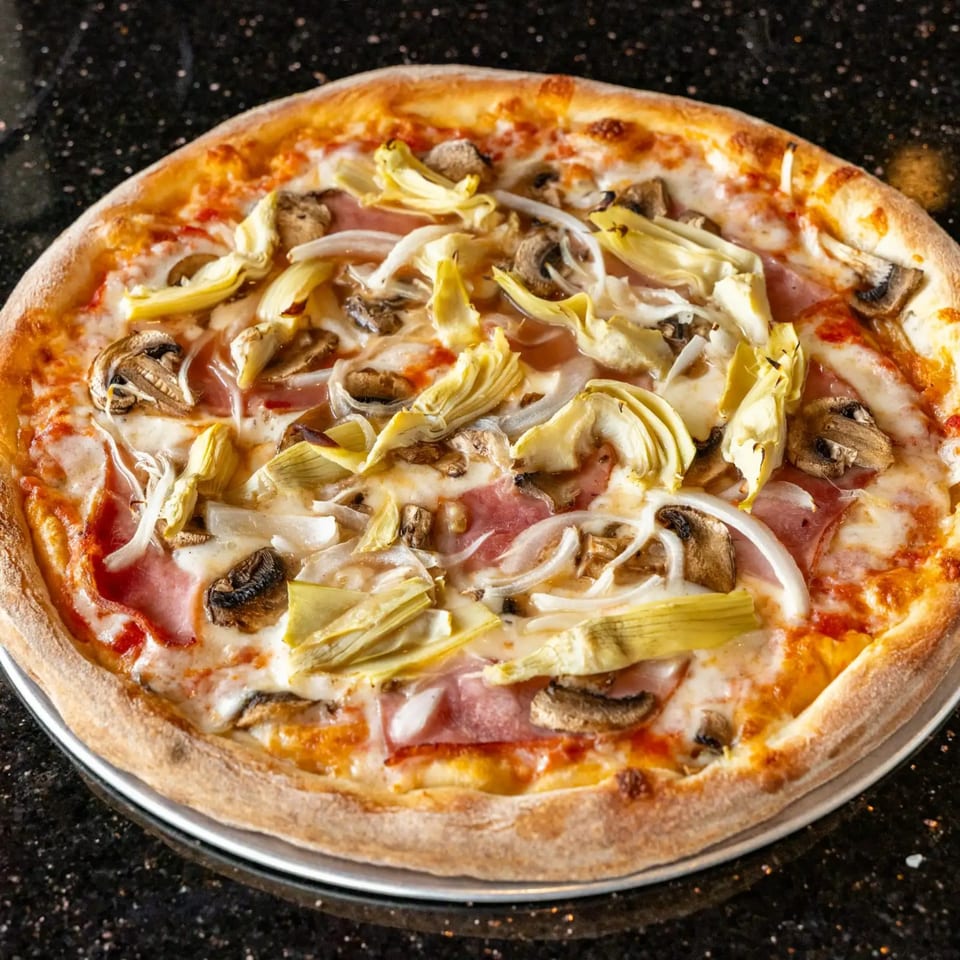 A whole pizza with ham, artichoke hearts, mushrooms, and onions on a dark speckled surface.