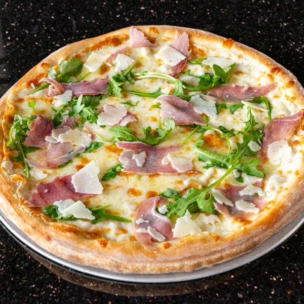 Grande Pizza's white pizza with prosciutto, arugula, and shaved Parmesan on a dark speckled surface.