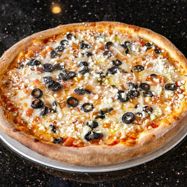 Grande Pizza's Greek pizza with black olives, feta cheese, and tomatoes on a dark speckled surface.