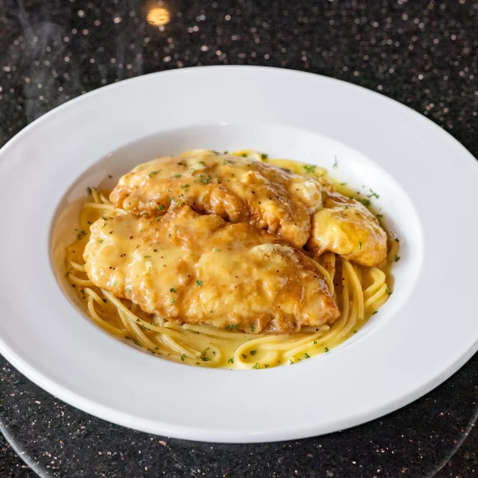 Chicken piccata with spaghetti in a white bowl, garnished with parsley, on a dark speckled surface.